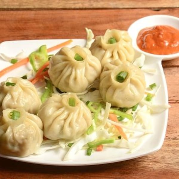 A plate of steamed momos, garnished with green onions, served with shredded vegetables and red dipping sauce.