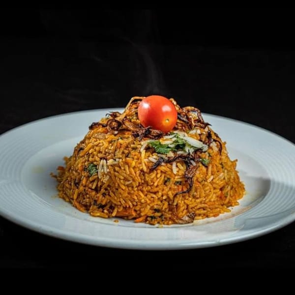 Steaming Indian biryani, garnished with a cherry tomato and crispy fried onions, on a white plate.