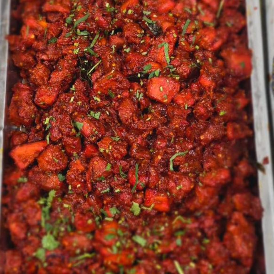 Close-up of vibrant red Indian chicken curry, garnished with fresh green herbs in a metal serving tray.