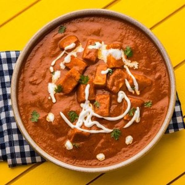 A bowl of Indian paneer butter masala, garnished with cream and cilantro, on a yellow table.